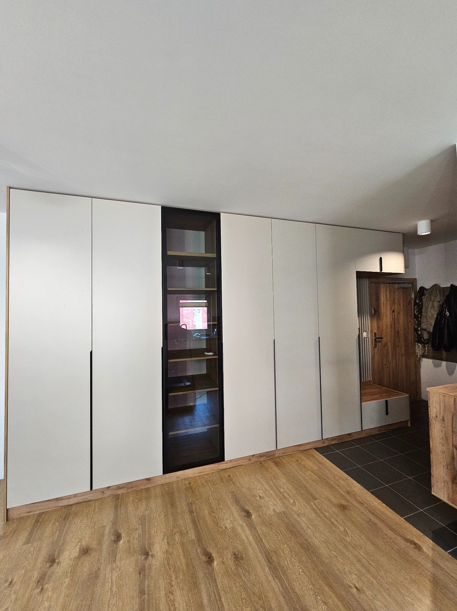 Biała szafa wnękowa z czarnymi uchwytami i przeszkloną witryną, dopasowana do drewnianej podłogi i drzwi wejściowych. Minimalistyczny design.