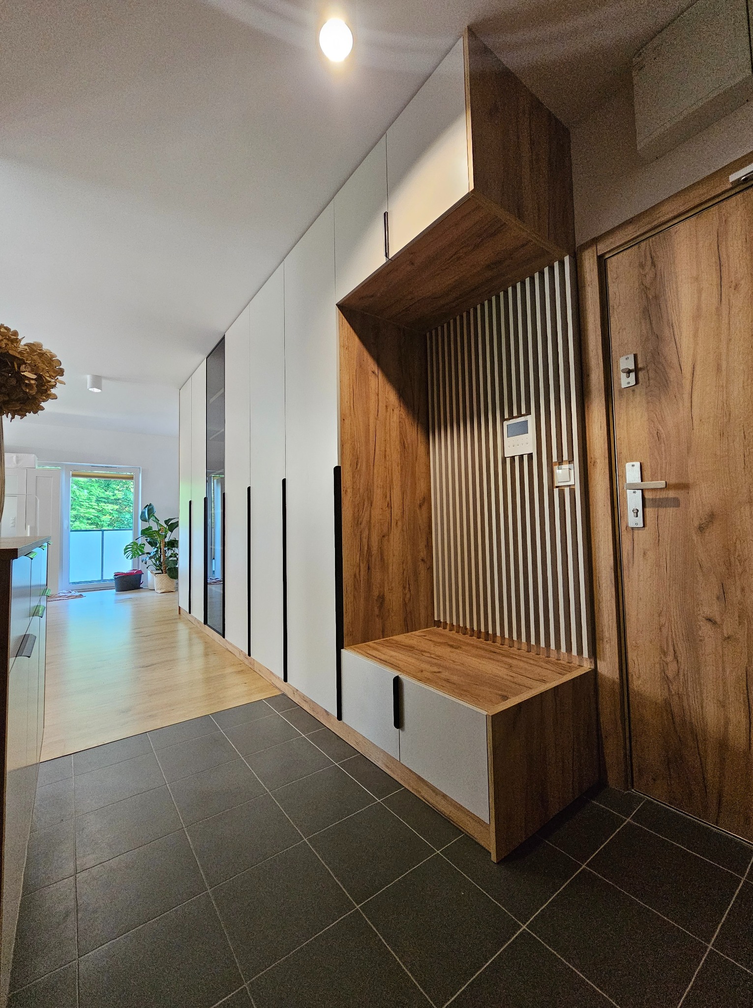 Nowoczesna zabudowa przedpokoju z szafami wnękowymi w bieli i drewnie, z siedziskiem i dekoracyjnymi lamelami. Minimalistyczny design, czarne uchwyty, szara podłoga.