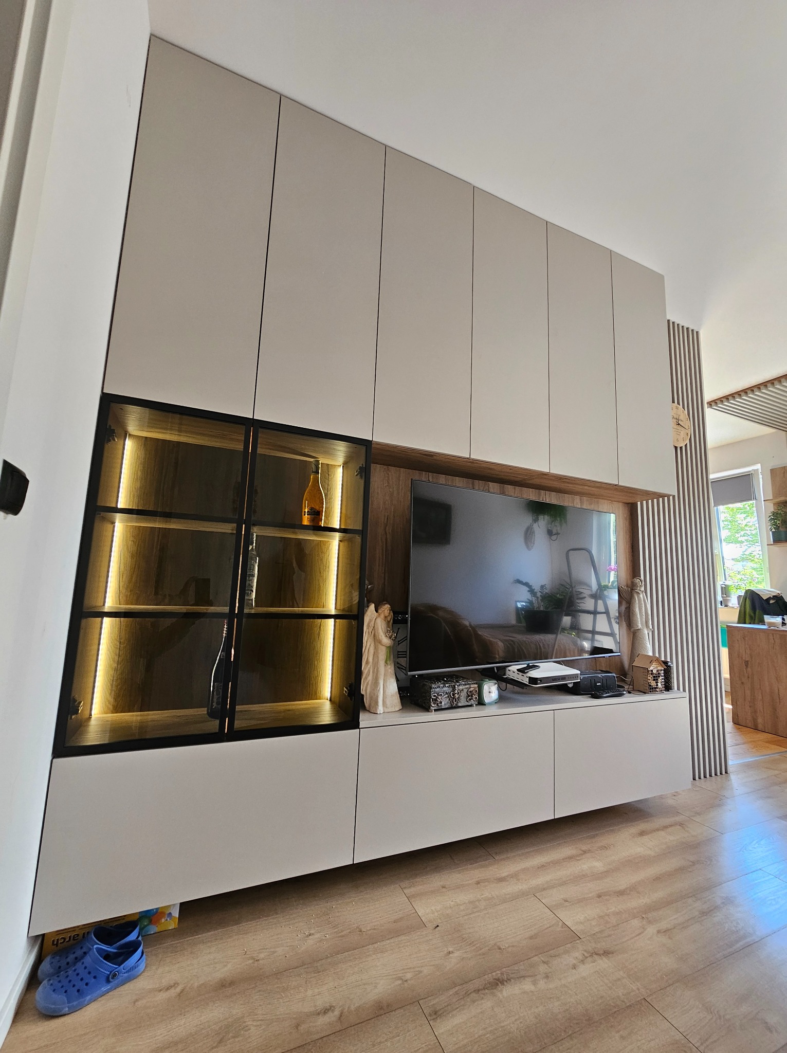 Nowoczesna zabudowa meblowa w salonie: szafki wiszące, przeszklona witryna z podświetleniem LED, szafka RTV z drewnianym akcentem. Ujęcie z dołu, widoczne panele podłogowe.