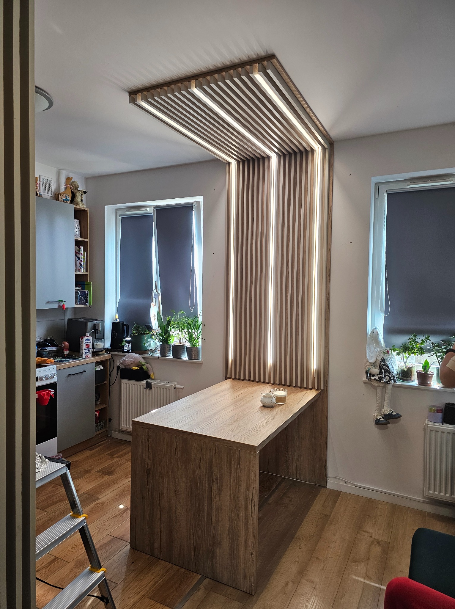 Nowoczesna kuchnia z drewnianym blatem i dekoracyjną ścianką z lameli z oświetleniem LED. Widoczne rośliny w oknach i drabina malarska.