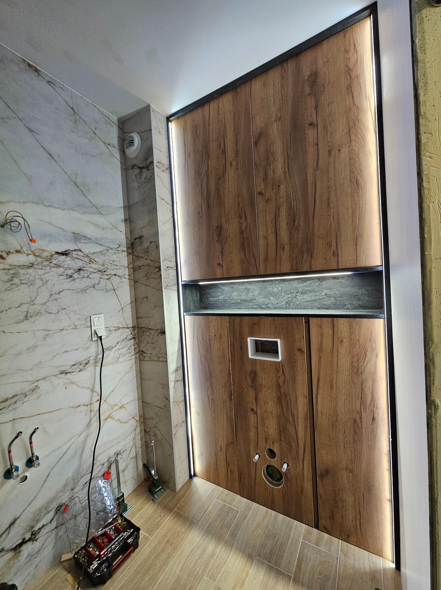 Nowoczesna zabudowa drewniana z podświetleniem LED w łazience, widoczne narzędzia i elementy instalacji, marmurowe wykończenie ściany.