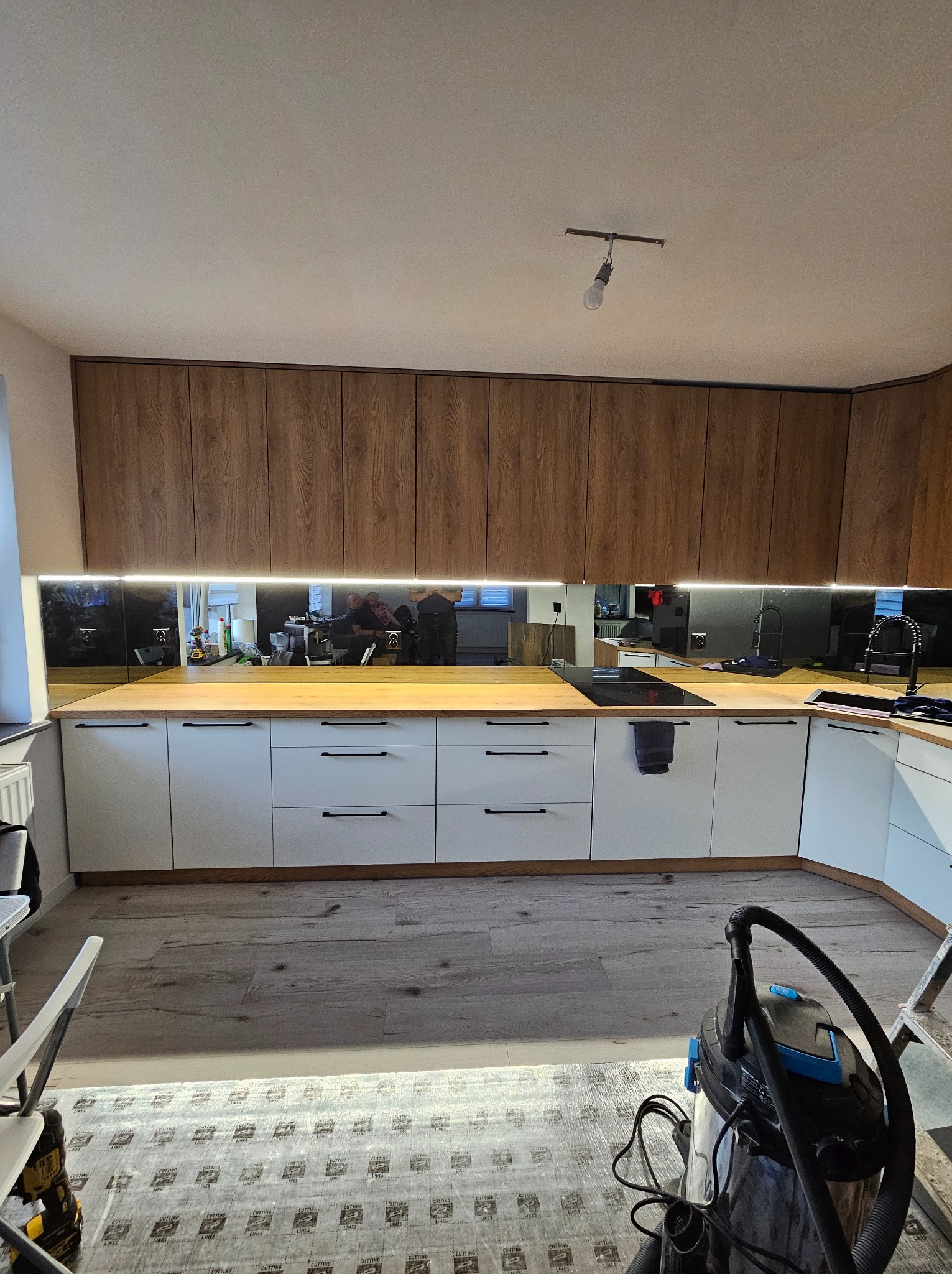 Nowoczesna kuchnia z białymi szafkami dolnymi i drewnianymi górnymi, oświetlona listwą LED pod szafkami. Na blacie płyta indukcyjna i czarny kran. Widoczny odkurzacz.