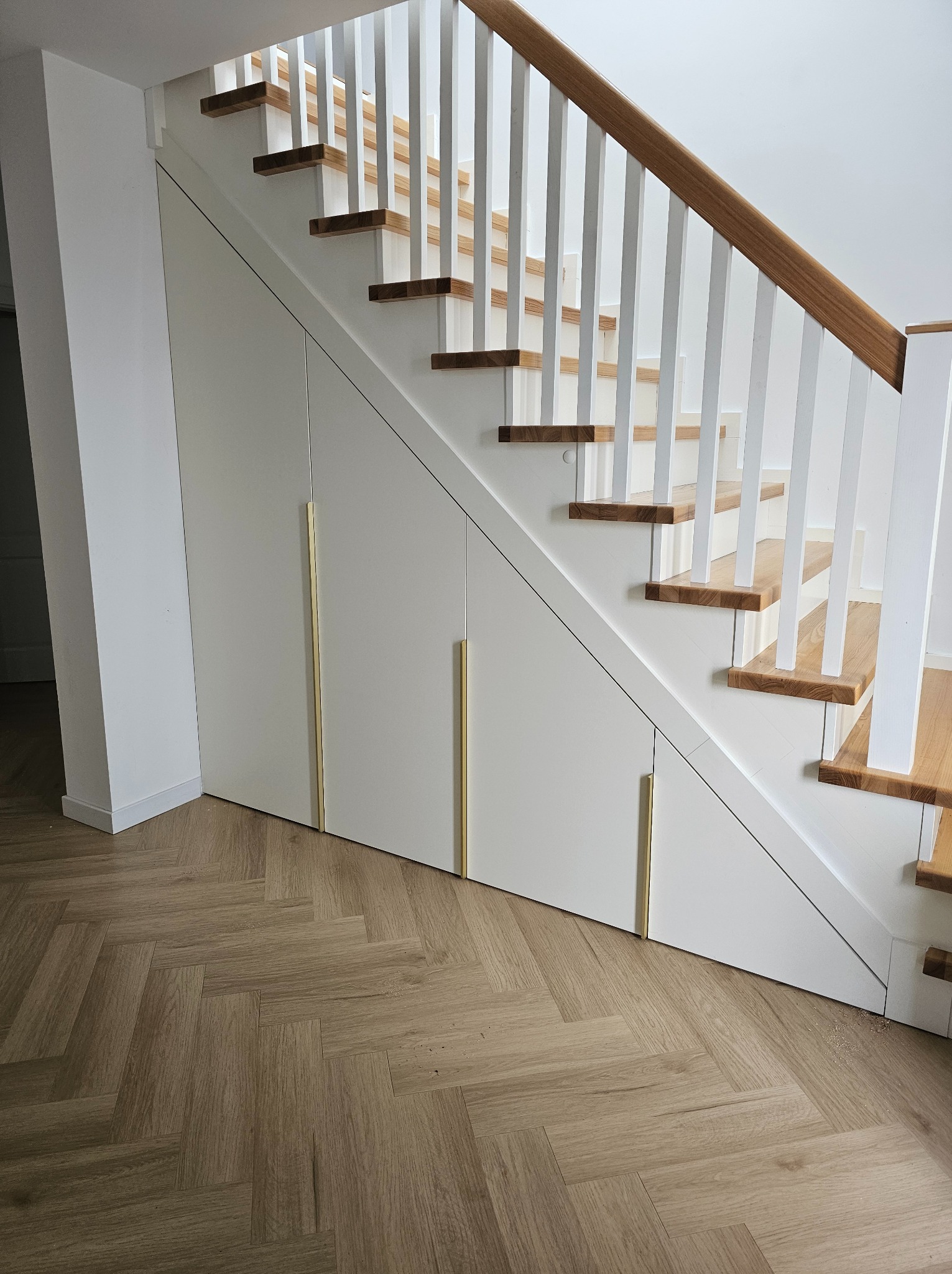 Białe schody z drewnianymi stopniami i szafkami pod schodami z długimi, złotymi uchwytami. Parkiet ułożony w jodełkę. Minimalistyczny design.