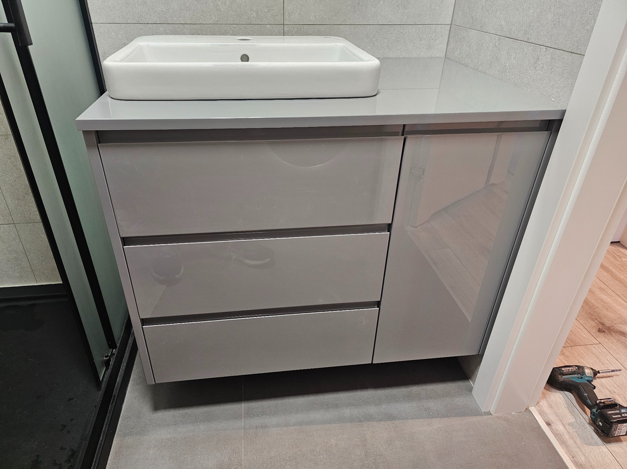 Szara szafka łazienkowa z umywalką, trzy szuflady i drzwiczki. Na podłodze wiertarka akumulatorowa. Minimalistyczny design, nowoczesne wykończenie.