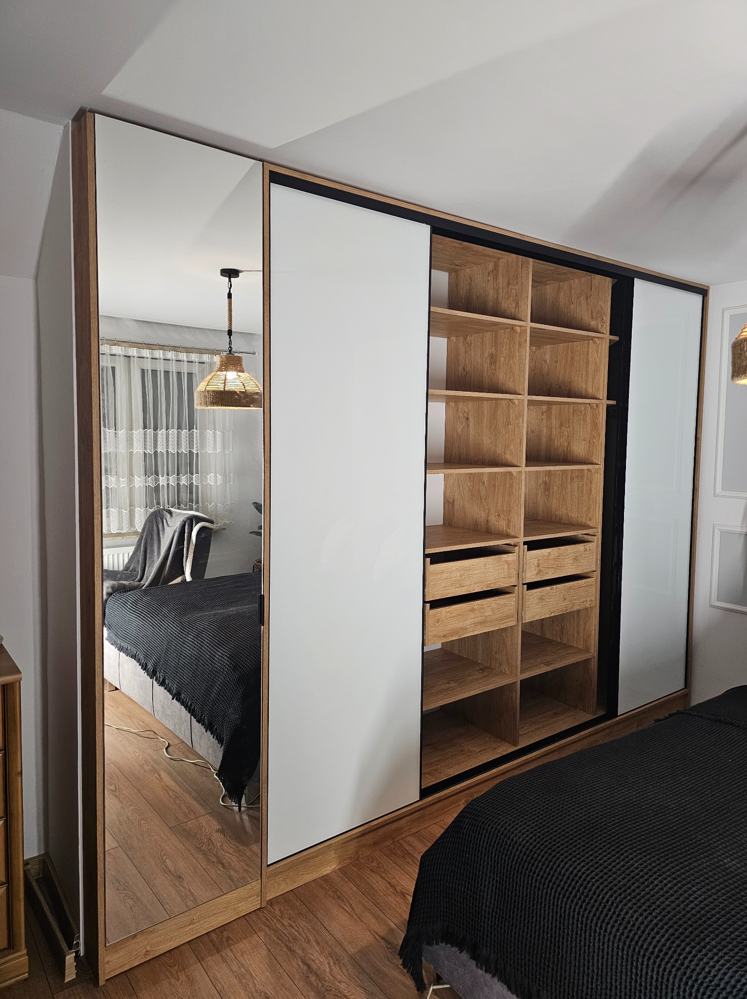 Szafa wnękowa z przesuwnymi drzwiami: lustrzane, białe i drewniane panele. Wnętrze z półkami i szufladami. Widok na sypialnię w lustrze.