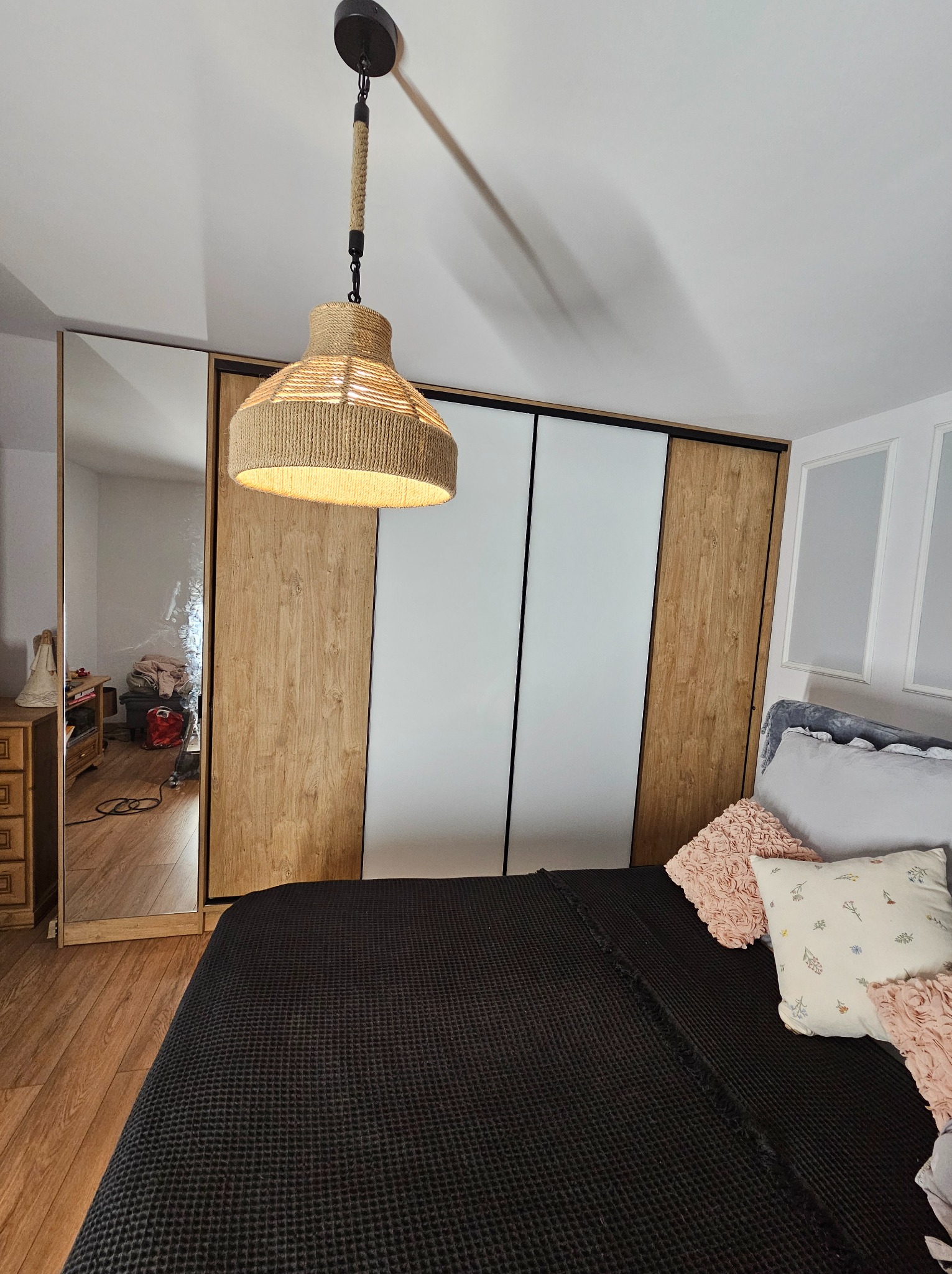 Szafa wnękowa z przesuwnymi drzwiami: drewniane panele po bokach, białe w środku. W tle widoczne łóżko i lampa z abażurem z naturalnego sznurka.
