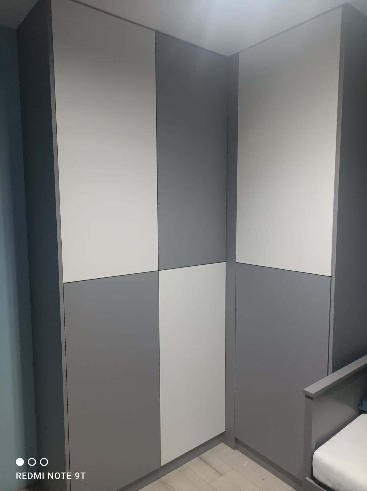 Nowoczesna szafa wnękowa z frontami w szachownicę, szaro-biała kolorystyka, minimalistyczny design, widoczny fragment dziecięcego łóżeczka, jasna podłoga.