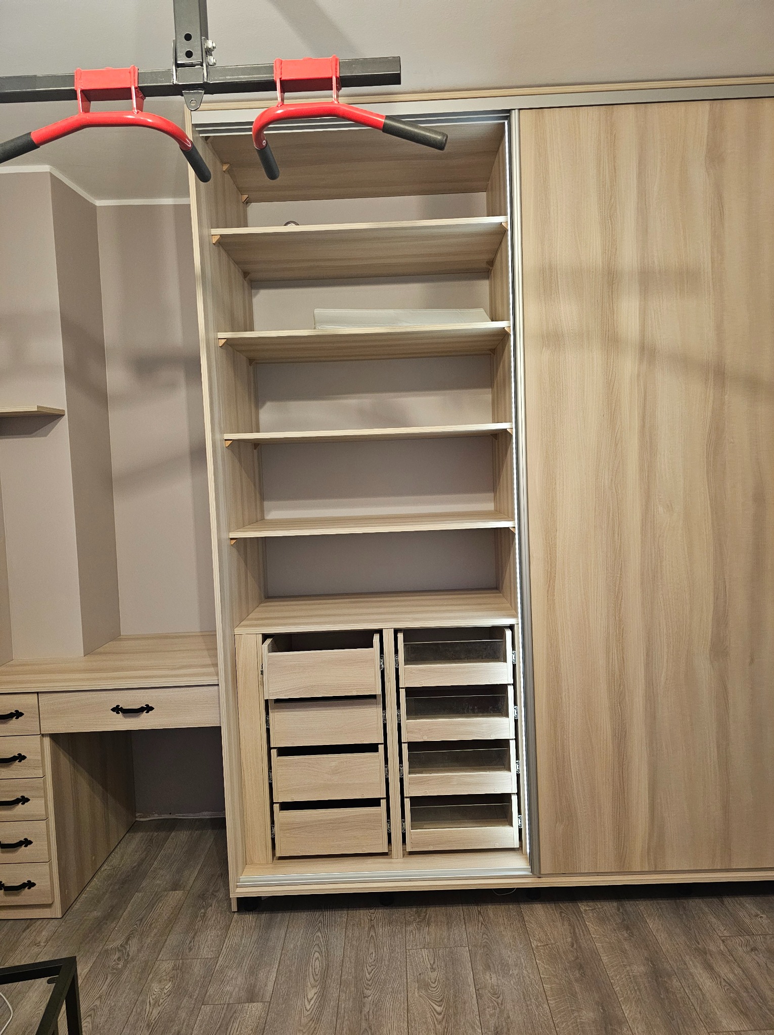 Jasna szafa wnękowa z przesuwnymi drzwiami i otwartymi półkami, uzupełniona biurkiem z szufladami. Nad szafą widoczny fragment drążka do podciągania.