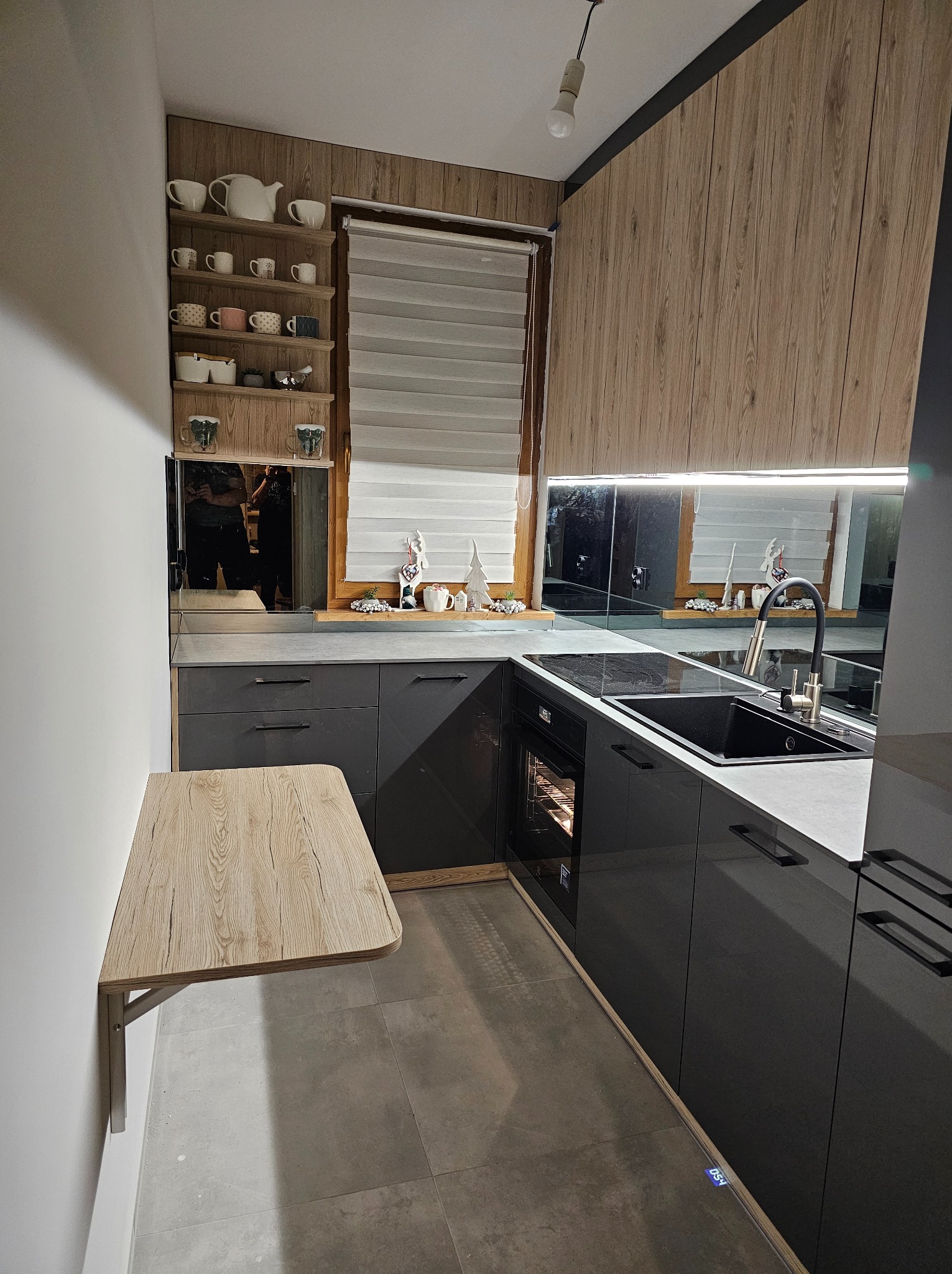 Nowoczesna kuchnia z szarymi szafkami, drewnianymi akcentami i rozkładanym blatem. Widoczne półki z dekoracjami i odbicie w lustrzanych panelach nad blatem.