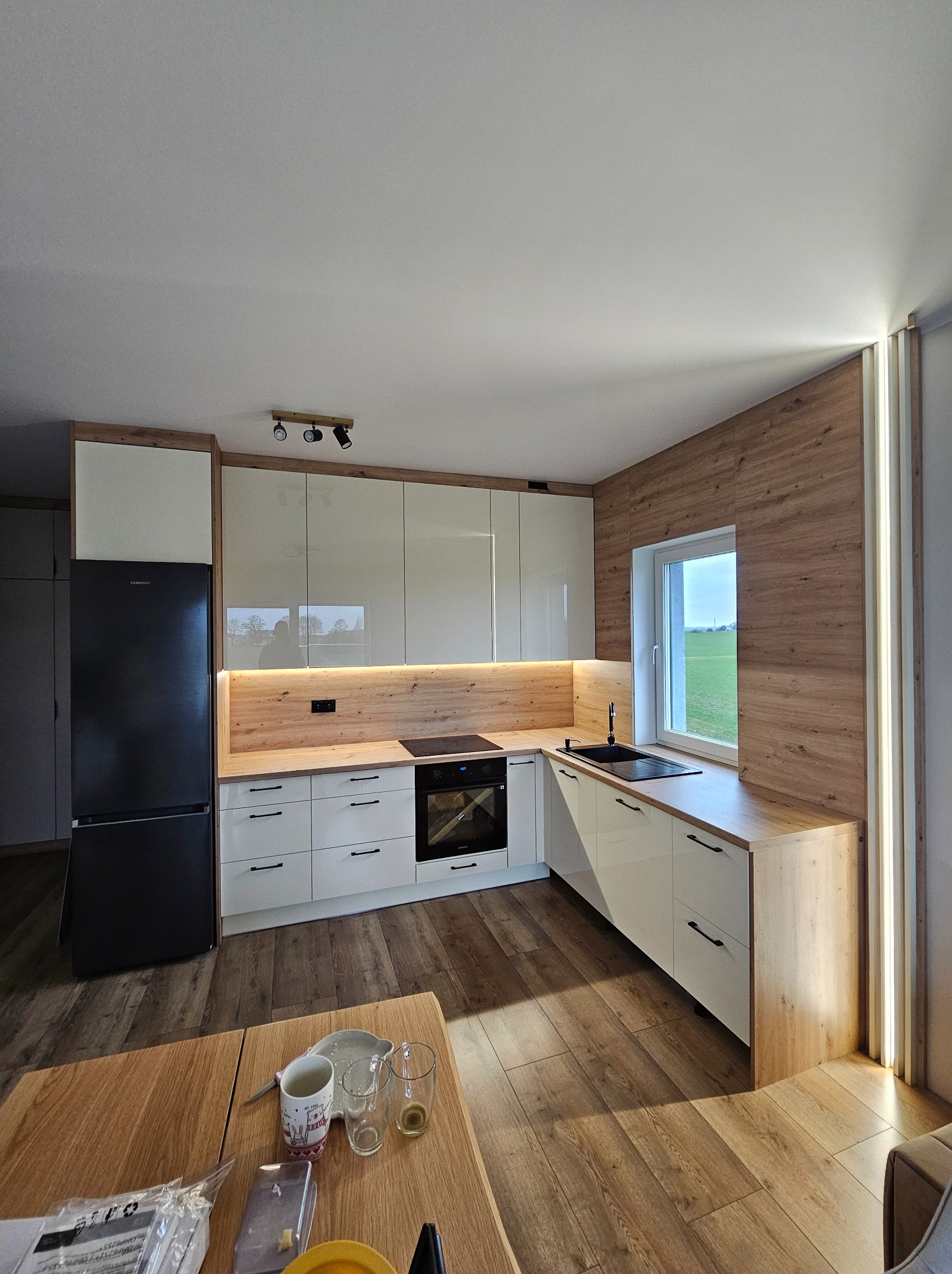 Nowoczesna kuchnia na wymiar z białymi frontami i drewnianymi blatami, oświetleniem LED pod szafkami i drewnianą ścianą. Widok na zielone pole przez okno.