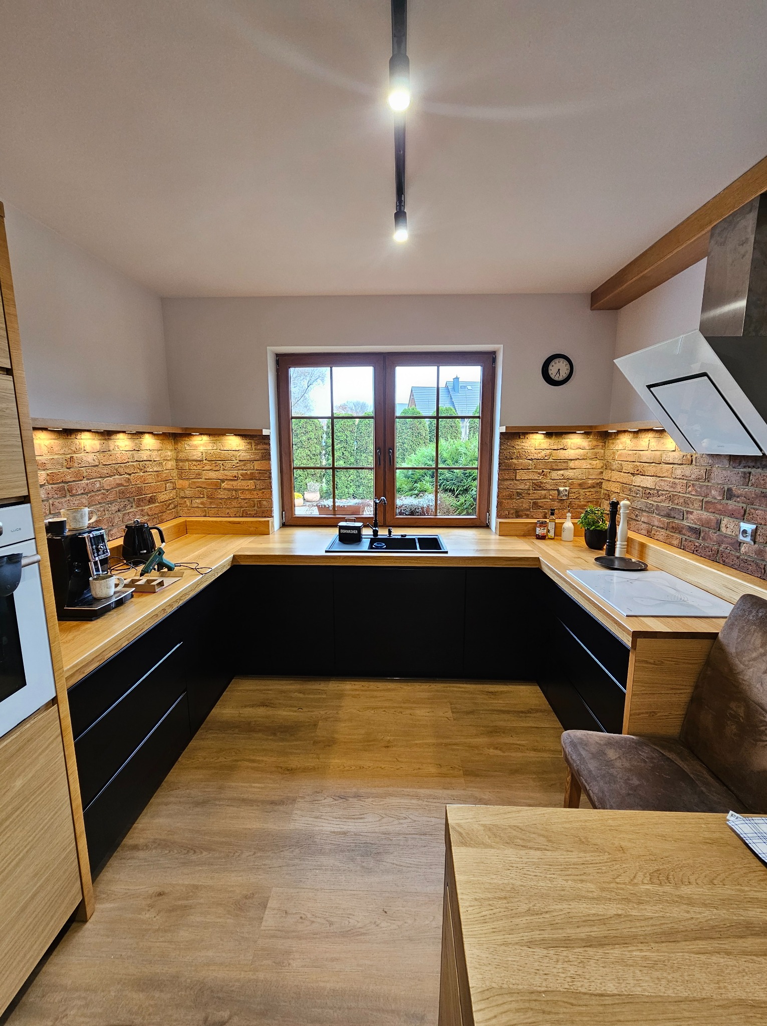 Nowoczesna kuchnia w kształcie litery U z czarnymi szafkami i drewnianymi blatami. Ceglana ściana nad blatem, okno z widokiem na ogród. Oświetlenie punktowe.