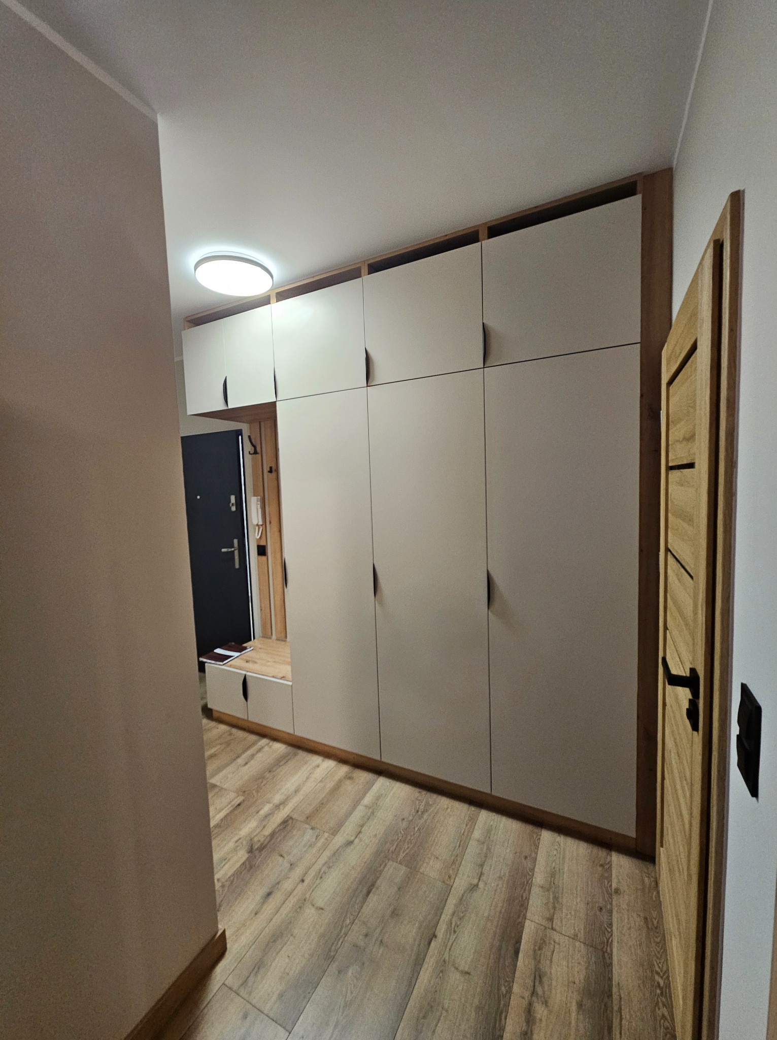Jasna szafa na wymiar z drewnianymi akcentami w przedpokoju. Minimalistyczny design, podłoga z paneli, drzwi wejściowe i wewnętrzne widoczne w tle.