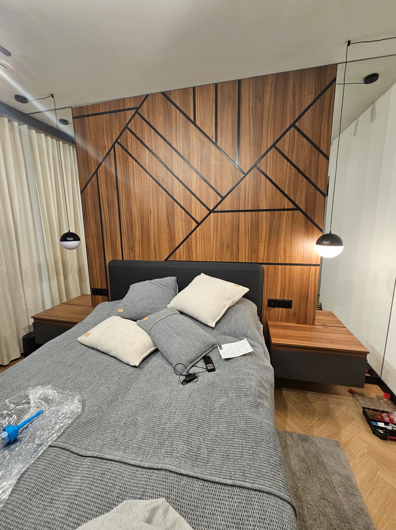 Nowoczesna sypialnia z łóżkiem, drewnianym panelem ściennym o geometrycznym wzorze i wiszącymi lampami. Widoczne szafki nocne z drewnianymi blatami i szara pościel.
