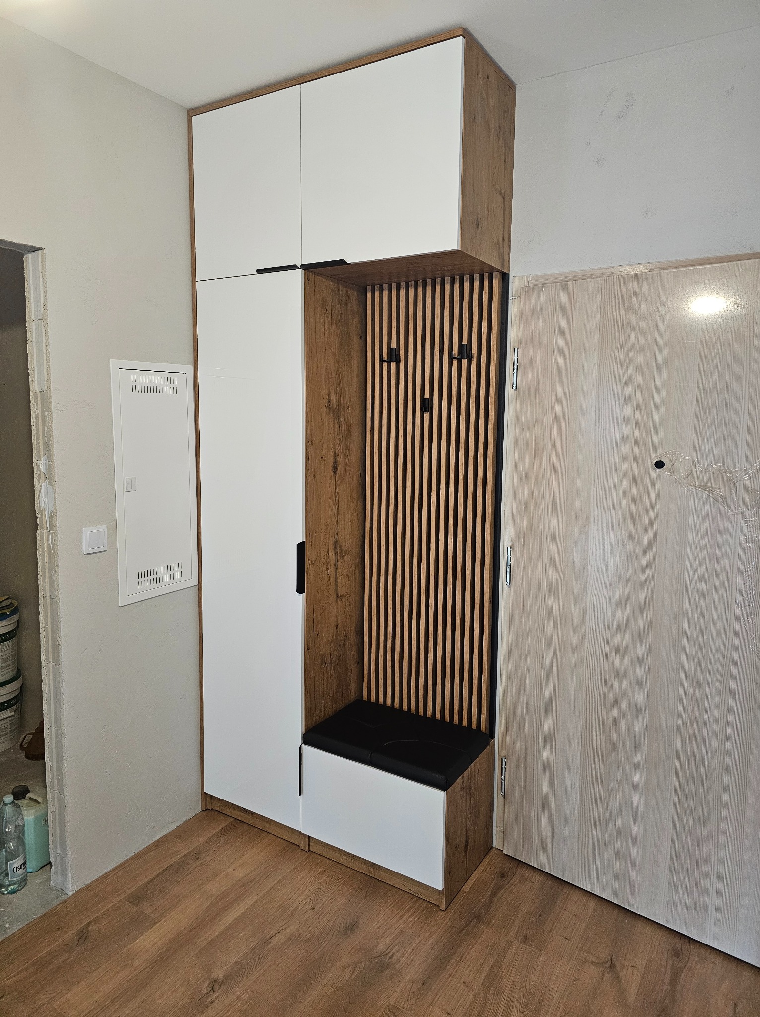 Funkcjonalna szafa wnękowa z siedziskiem i wieszakami, wykończona w bieli i drewnie, idealna do przedpokoju. Nowoczesny design z lamelami i czarnymi uchwytami.