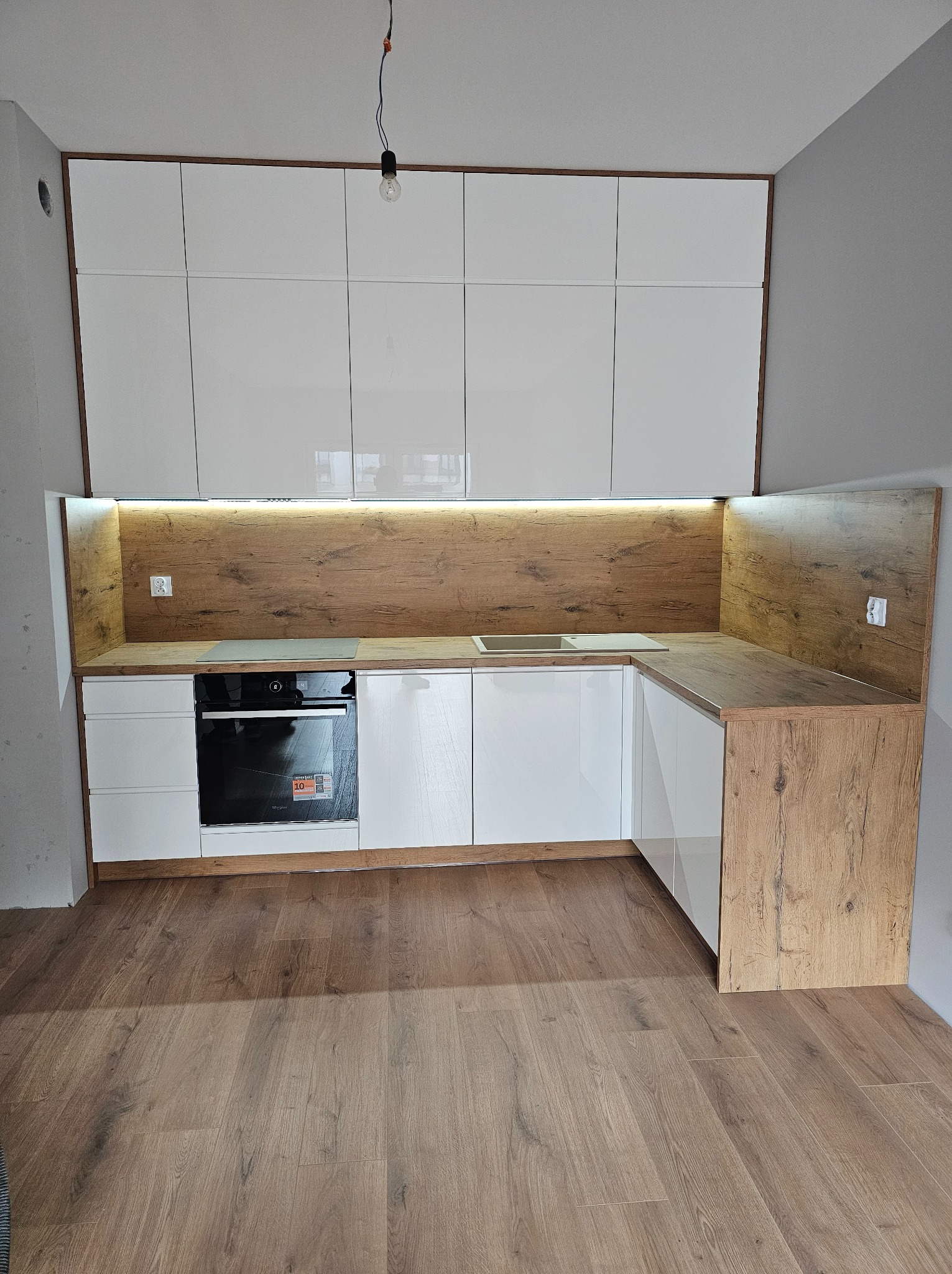 Nowoczesna kuchnia w kształcie litery L z białymi, błyszczącymi frontami i drewnianym blatem. Oświetlenie LED pod szafkami górnymi. Widoczny piekarnik i drewniana podłoga.