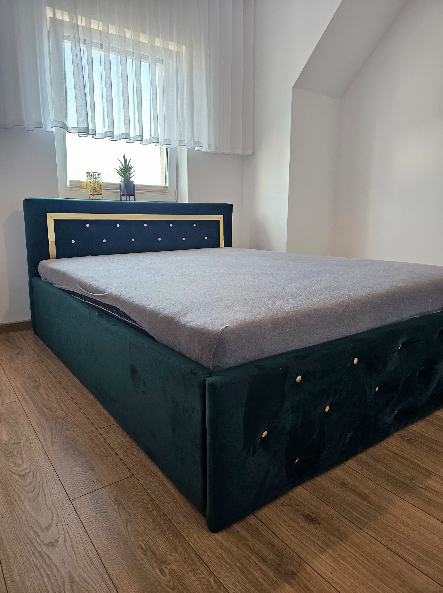 Ciemnozielone, tapicerowane łóżko z pikowanym wezgłowiem i złotymi detalami, ustawione na drewnianej podłodze w jasnym pokoju z oknem.