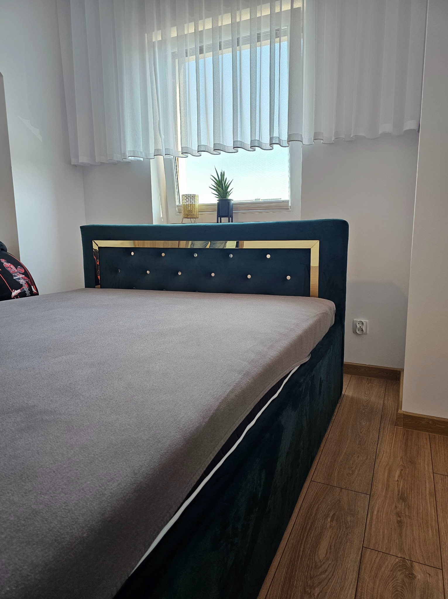 Ciemnozielone łóżko tapicerowane z pikowanym zagłówkiem i złotymi detalami, ustawione na drewnianej podłodze, z oknem w tle.