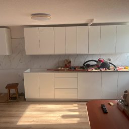 Krystian Beeger - Montaż białych szafek kuchennych z marmurowym backsplashiem. Na blacie leżą narzędzia, a obok stoi taboret. Widoczny parkiet i okno z grzejnikiem.