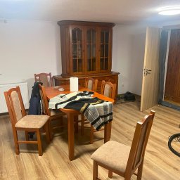 Krystian Beeger - Jasne wnętrze jadalni z drewnianym stołem, krzesłami i kredensem. Na stole leży pled. Widoczna podłoga z paneli i białe ściany. W tle otwarte drzwi.