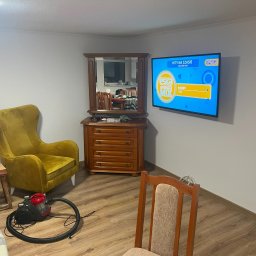 Krystian Beeger - Umeblowane wnętrze pokoju z drewnianą komodą z lustrem, telewizorem na ścianie, fotelami i odkurzaczem na parkiecie. Widok na odnowione, jasne pomieszczenie.