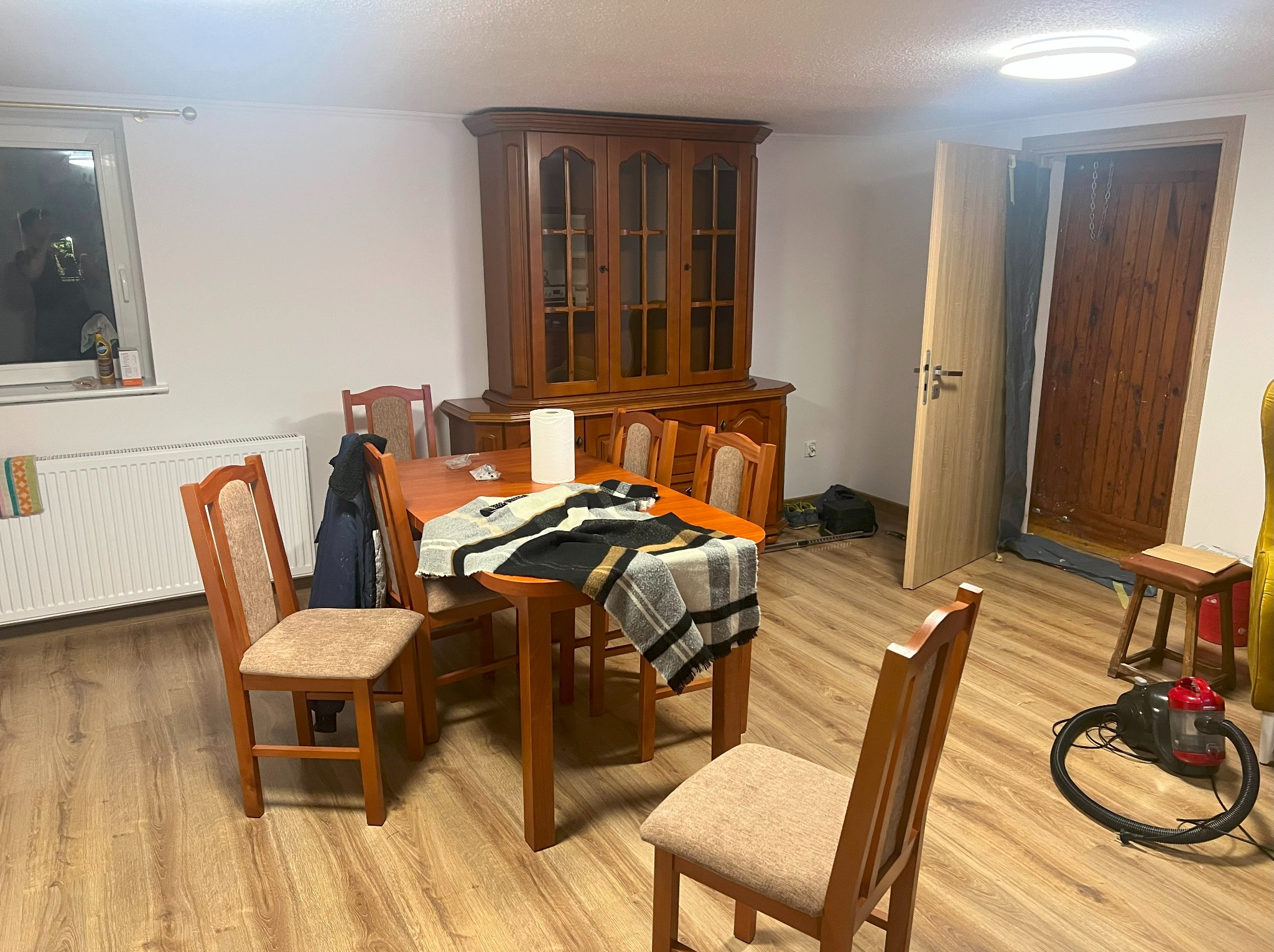 Jasne wnętrze jadalni z drewnianym stołem, krzesłami i kredensem. Na stole leży pled. Widoczna podłoga z paneli i białe ściany. W tle otwarte drzwi.