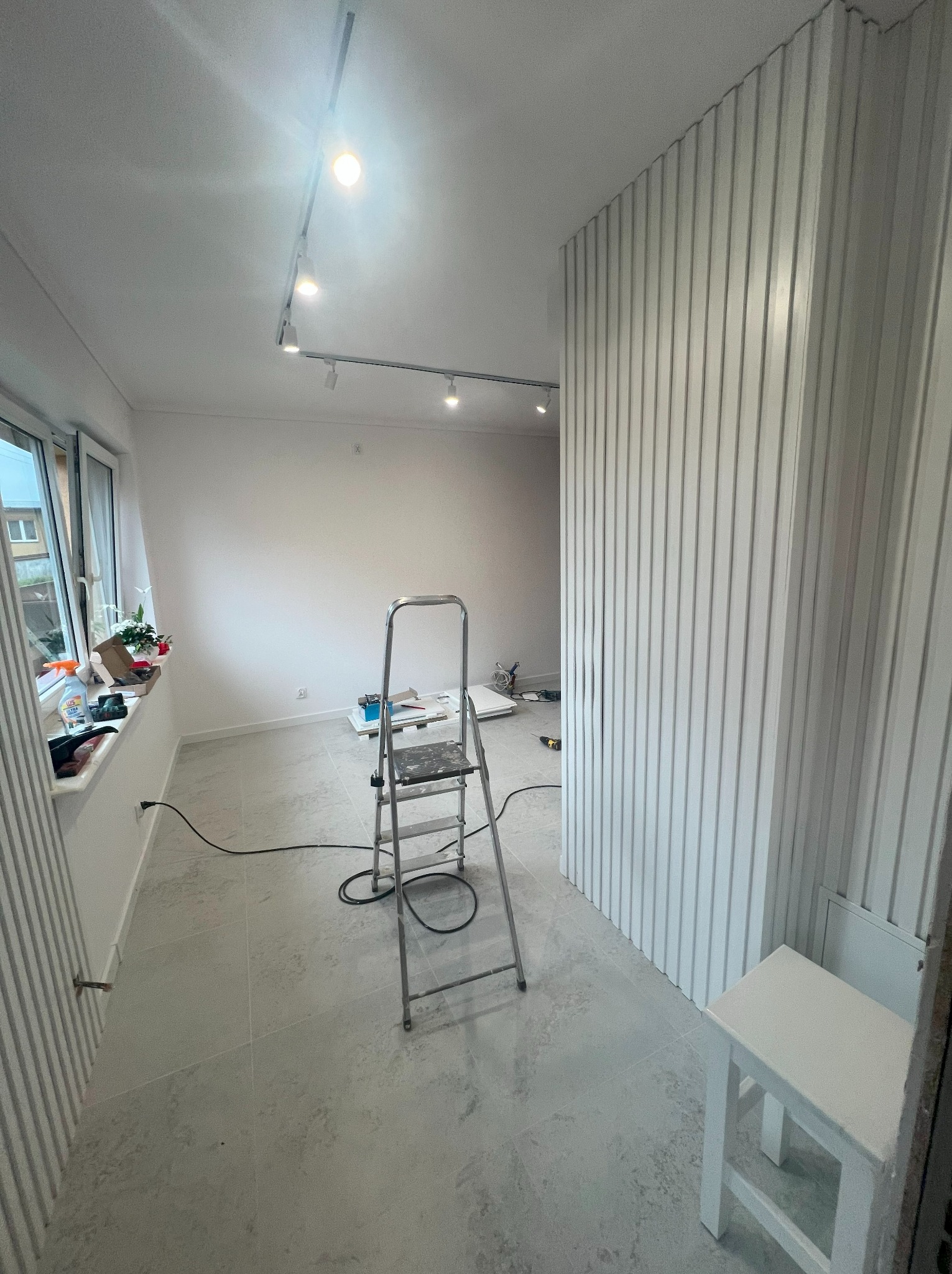 Jasne wnętrze w trakcie wykańczania: białe ściany z dekoracyjnymi panelami, drabina, widoczne narzędzia i akcesoria malarskie przy oknie. Oświetlenie szynowe.