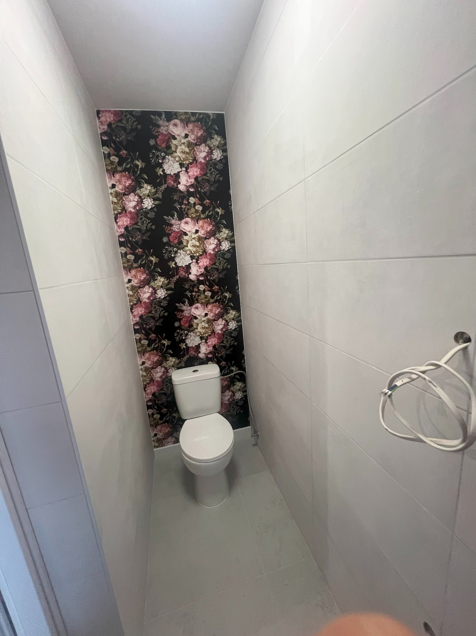 Wąska toaleta po remoncie: białe płytki, ściana z motywem kwiatowym, podwieszana muszla WC. Widoczne przewody na ścianie.
