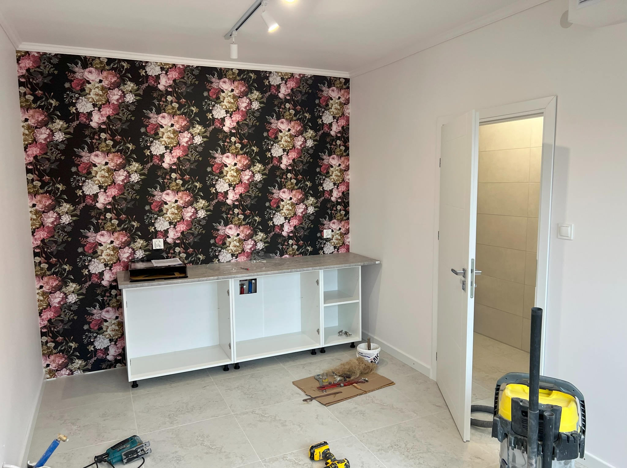 Wnętrze kuchni w trakcie montażu: szafki z blatem na tle ściany w kwiaty, narzędzia na podłodze, otwarte drzwi do łazienki w tle. Widoczne oświetlenie sufitowe.