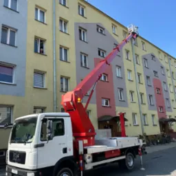 Podnośnik koszowy na tle bloku z kolorową elewacją. W oknie widać osobę obserwującą prace. Czerwone ramię podnośnika sięga do górnych pięter. Słoneczny dzień.