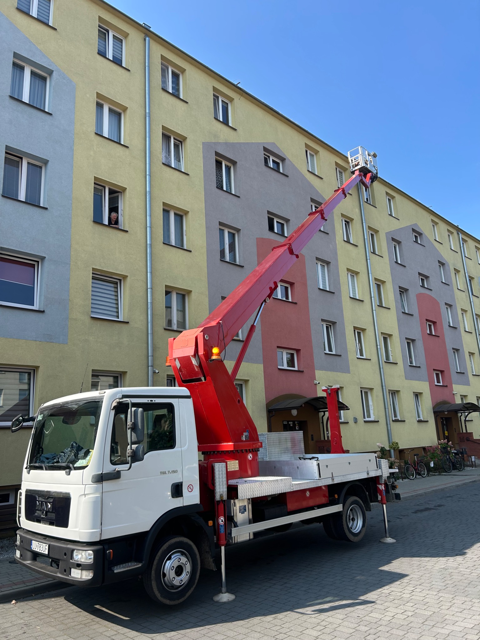 Podnośnik koszowy na tle bloku z kolorową elewacją. W oknie widać osobę obserwującą prace. Czerwone ramię podnośnika sięga do górnych pięter. Słoneczny dzień.