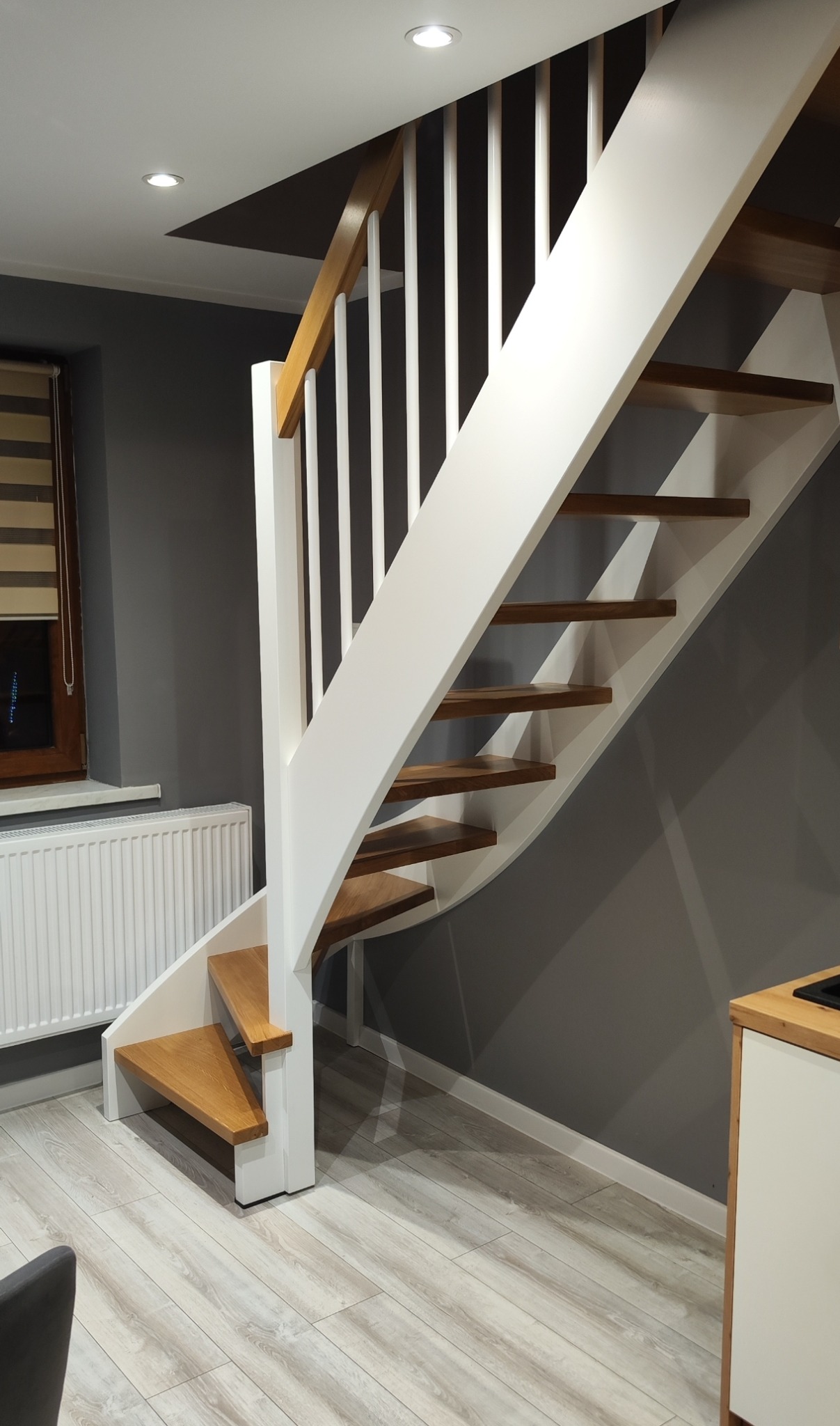 Nowoczesne schody drewniane z białymi balustradami i drewnianymi stopniami, widoczne w jasnym wnętrzu z szarą ścianą i grzejnikiem pod oknem.