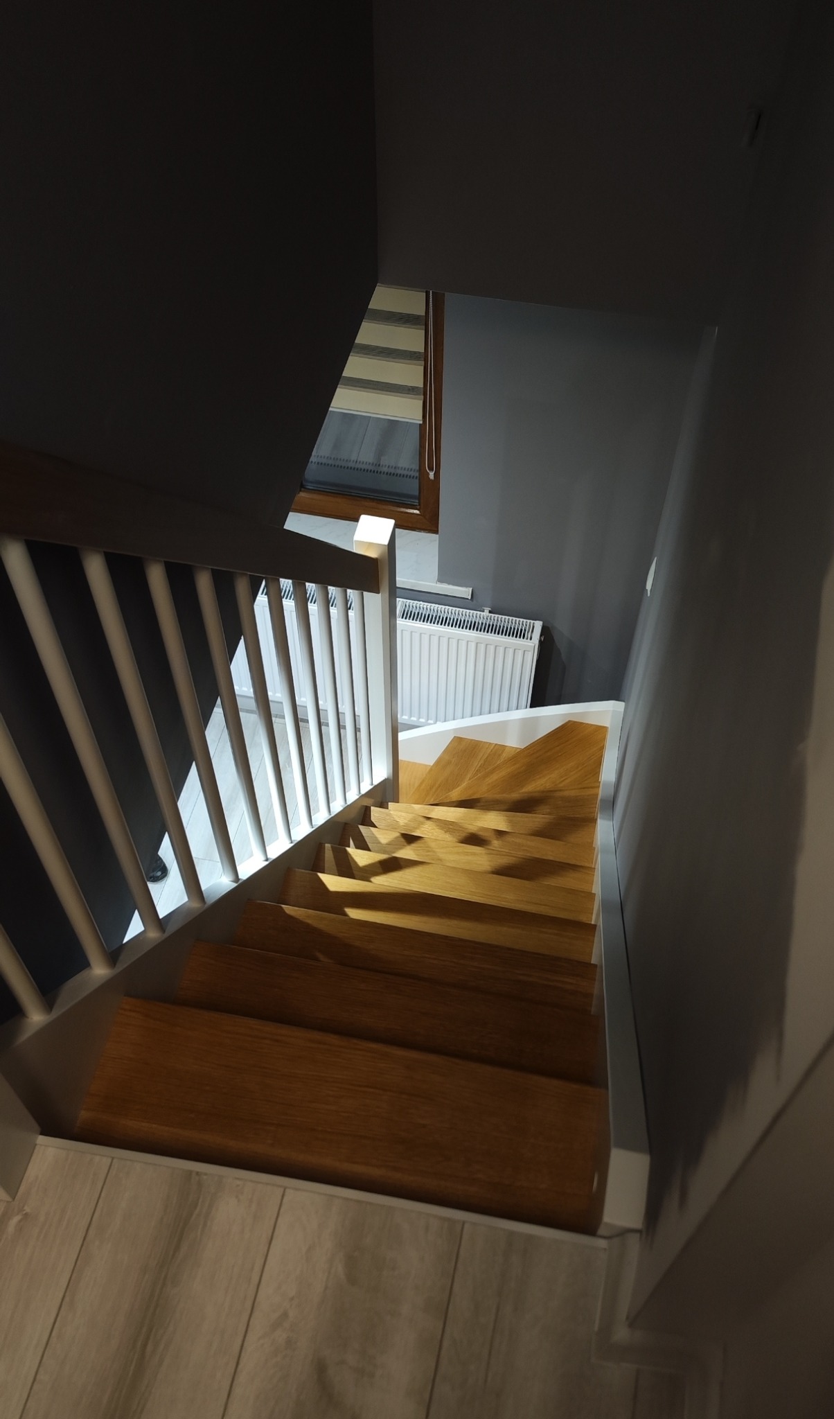 Drewniane schody z białymi balustradami w nowoczesnym wnętrzu. Widok z góry na stopnie o ciepłym odcieniu drewna i szare ściany. Okno z żaluzjami w tle.