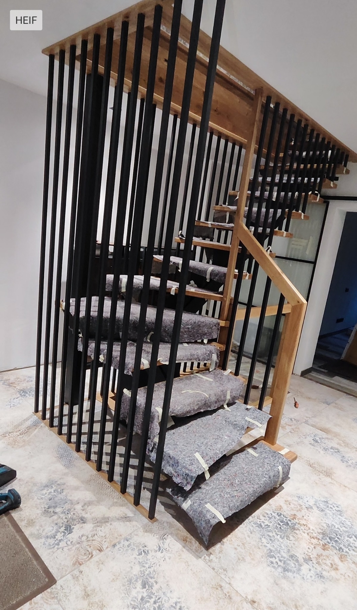 Nowoczesne schody drewniane z czarnymi, pionowymi balustradami, zabezpieczone na czas remontu. Widok z dołu, podkreślający minimalistyczny design i jasne drewno.