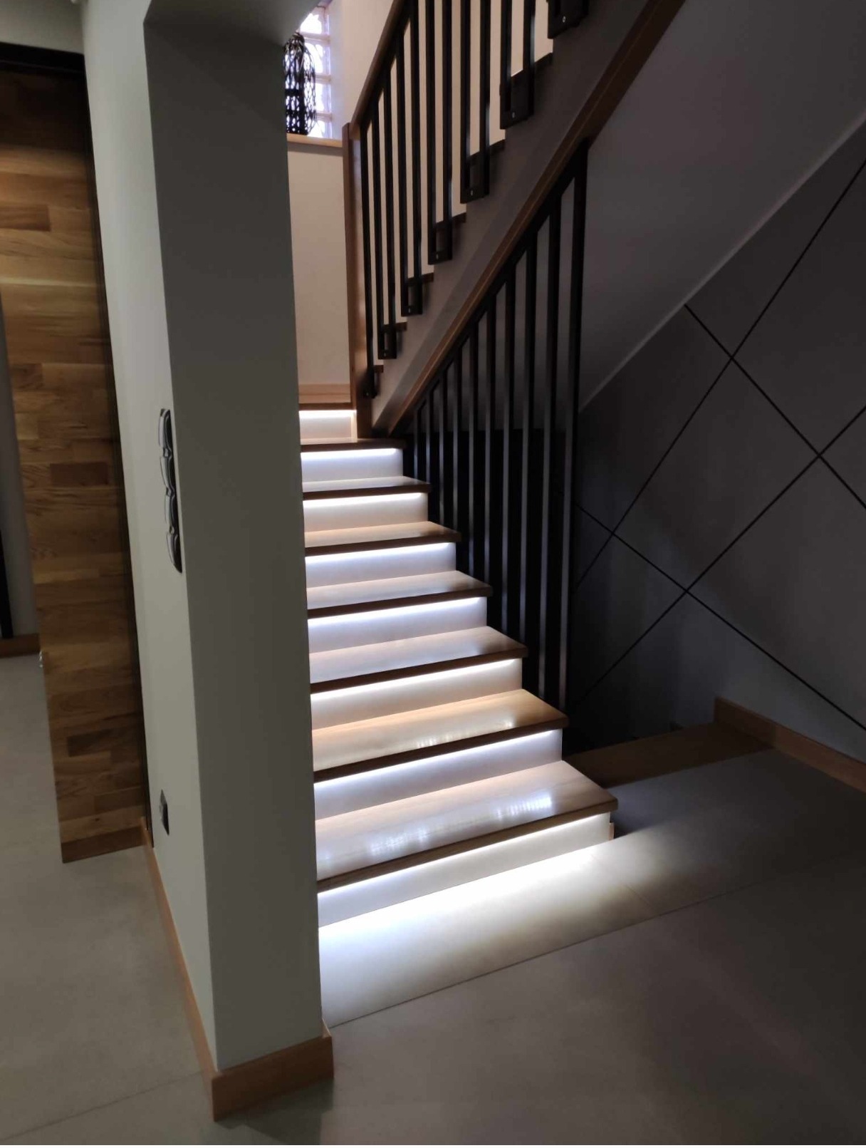 Nowoczesne schody drewniane z podświetleniem LED, czarne balustrady, szara ściana z geometrycznym wzorem. Minimalistyczny design, ciepłe światło.