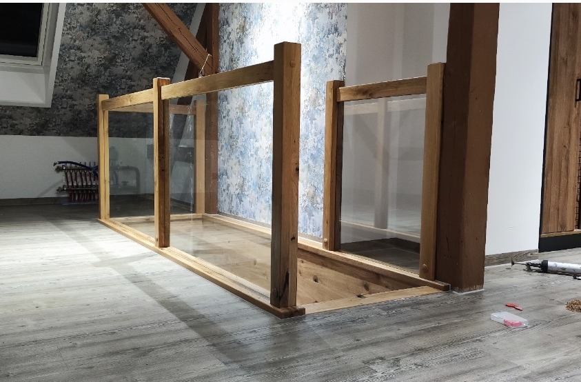 Drewniana konstrukcja z szybami, prawdopodobnie balustrada schodów, w surowym stanie, widoczne narzędzia i elementy montażowe. Ujęcie z niskiej perspektywy.