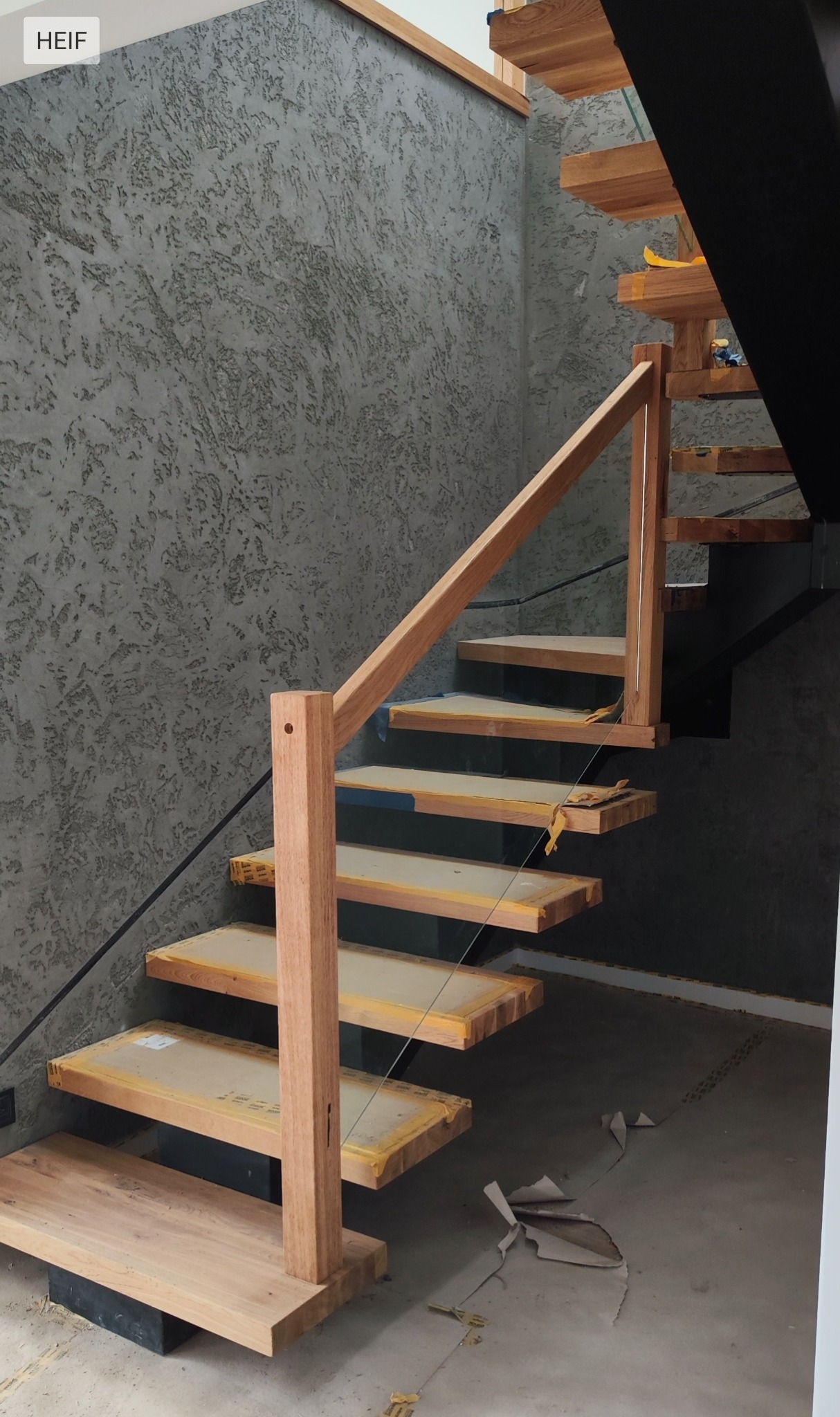 Nowoczesne schody drewniane z balustradą ze szkła, widok z dołu. Ściana z szarą, teksturowaną farbą. Elementy zabezpieczone folią ochronną.