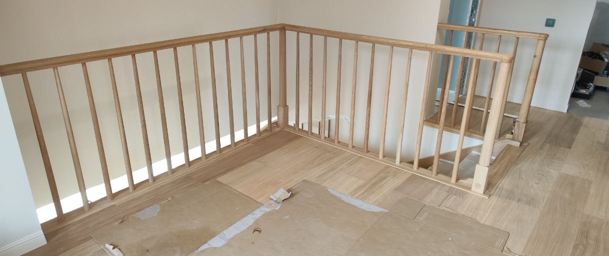 Jasne, drewniane balustrady schodowe z okrągłymi szczeblami, zamontowane na drewnianej podłodze. Widoczne zabezpieczenia podłogi. Ujęcie z góry.