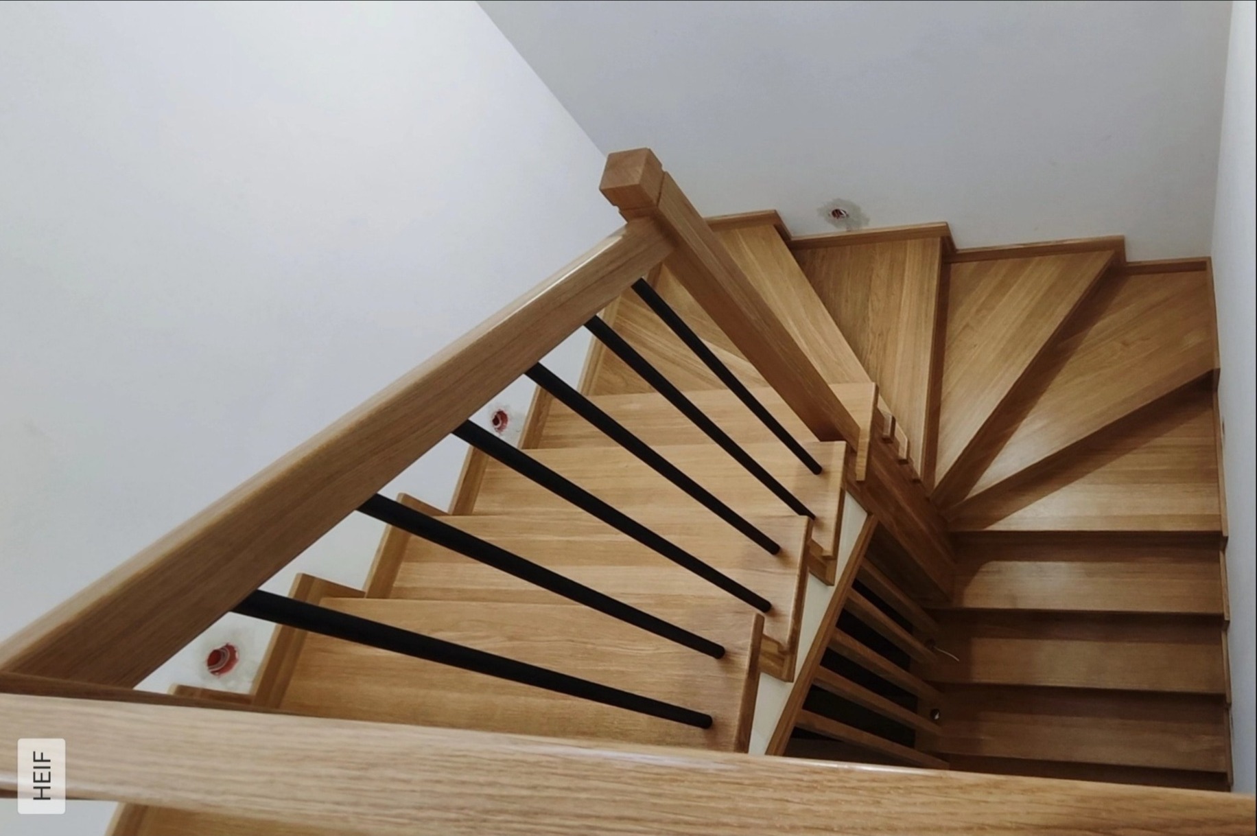 Drewniane schody z czarnymi balustradami, widok z góry, ukazujący zakręt schodów. Jasne drewno i minimalistyczny design.