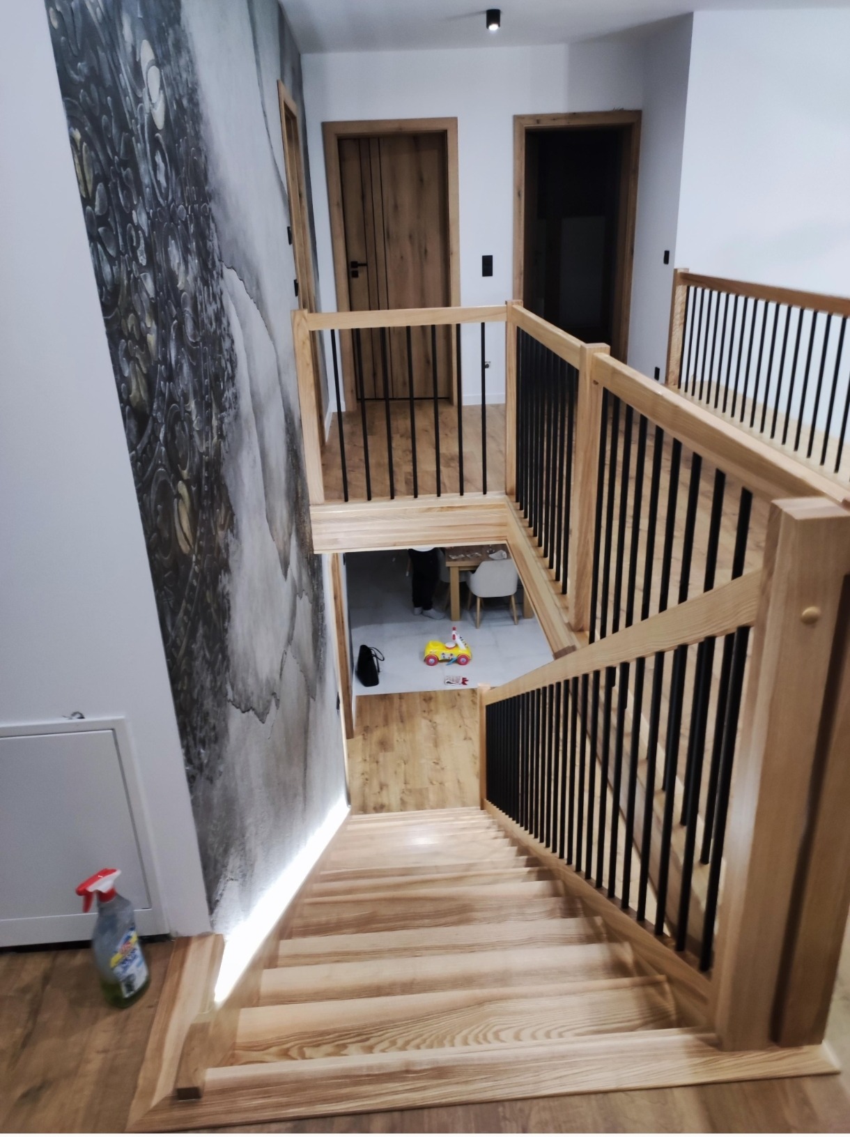 Jasne, drewniane schody z czarnymi balustradami, oświetlone od dołu, prowadzące w dół. Na ścianie dekoracyjna tapeta. Widok z góry.