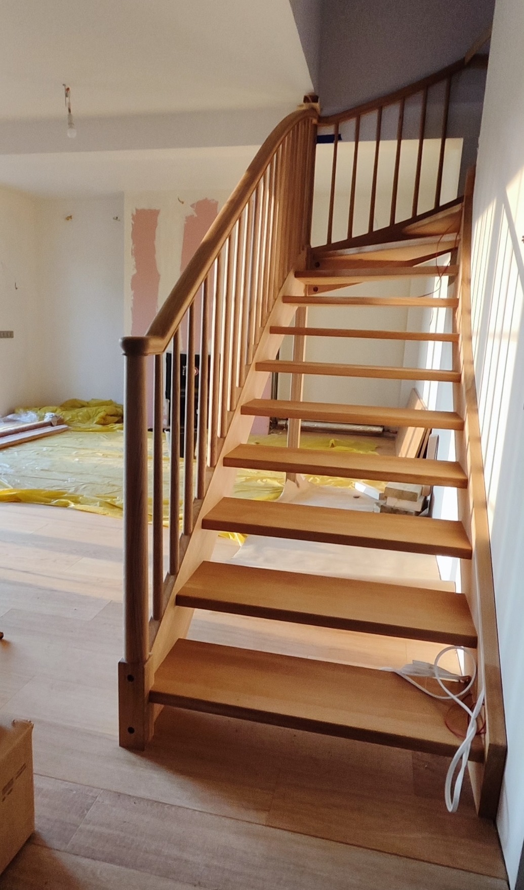 Jasne, drewniane schody z poręczą, zakręcające w prawo, w nowym domu. W tle widoczne zabezpieczenia podłogi folią malarską i ślady po pracach wykończeniowych.