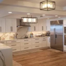 Nowoczesna kuchnia z białymi szafkami, marmurowym blatem i drewnianą podłogą. Oświetlenie punktowe i dekoracyjne lampy wiszące.