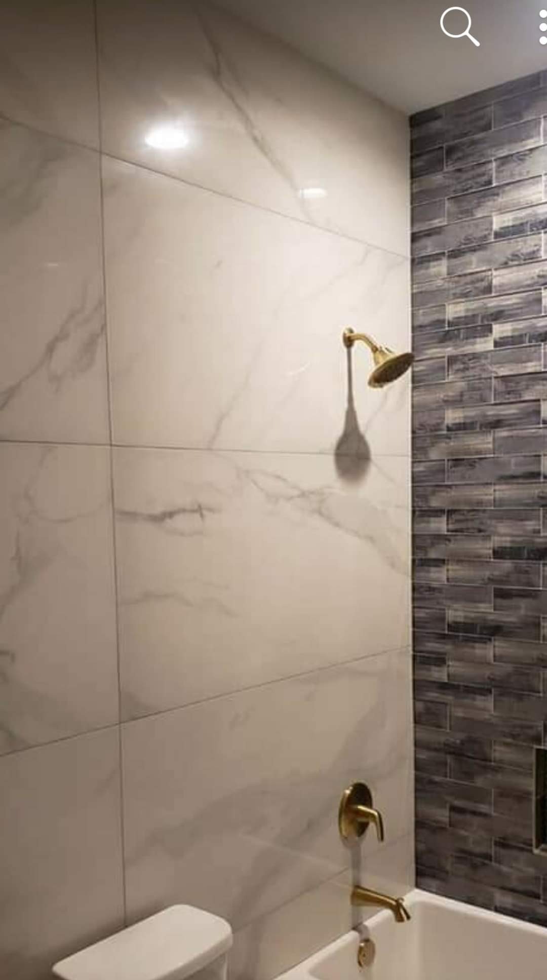 Wykończona łazienka z białymi płytkami imitującymi marmur i szarymi, wąskimi płytkami. Złota armatura, bateria i deszczownica. Widoczna toaleta i wanna.