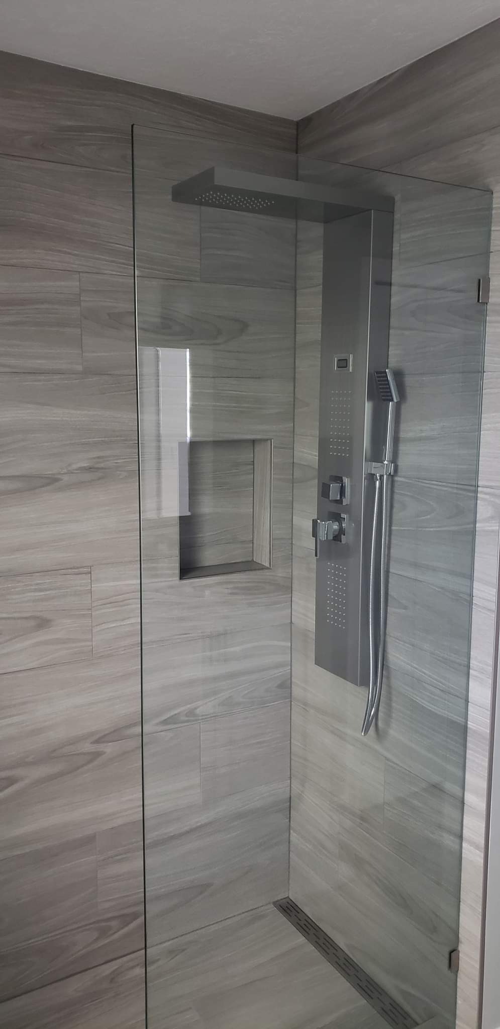 Nowoczesna kabina prysznicowa z szarymi płytkami imitującymi drewno i panelem natryskowym, widoczna kratka odpływowa w podłodze.