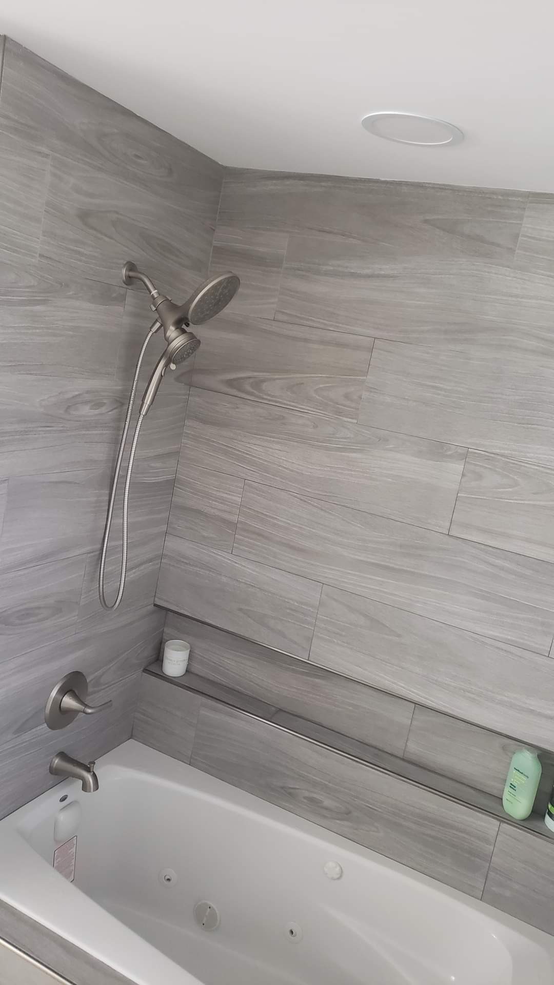 Wykończona łazienka z wanną jacuzzi, szarą glazurą imitującą drewno i półką na kosmetyki. Bateria prysznicowa w kolorze szczotkowanego niklu.