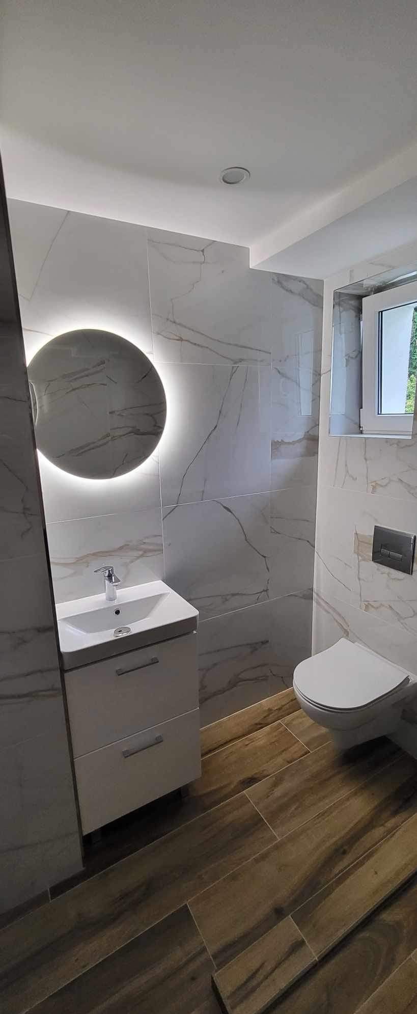 Nowoczesna łazienka po remoncie: białe marmurowe płytki, okrągłe lustro z podświetleniem LED, drewnopodobna podłoga, wisząca toaleta i biała szafka z umywalką.