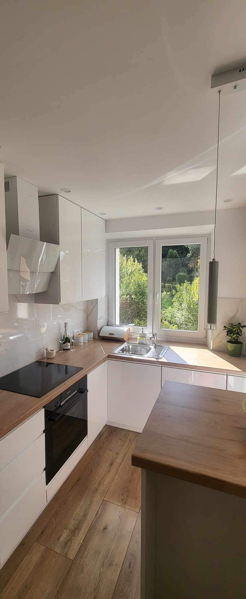 Jasna kuchnia z drewnianym blatem i podłogą. Widok na zieleń za oknem. Nowoczesne białe szafki i sprzęt AGD. Minimalistyczna lampa wisząca.