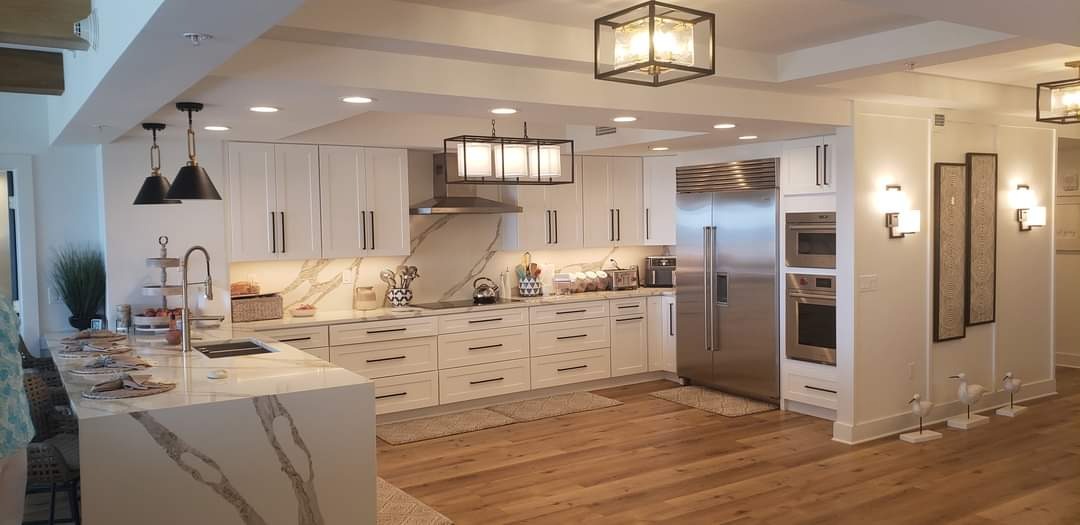 Nowoczesna kuchnia z białymi szafkami, marmurowym blatem i drewnianą podłogą. Oświetlenie punktowe i dekoracyjne lampy wiszące.