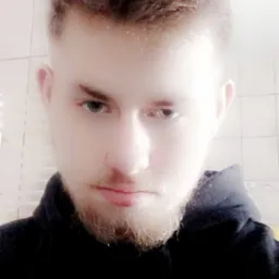 Portret młodego mężczyzny z lekkim zarostem, skierowany w dół, z intensywnym spojrzeniem na tle białych płytek. Ujęcie z góry, oświetlenie rozproszone.