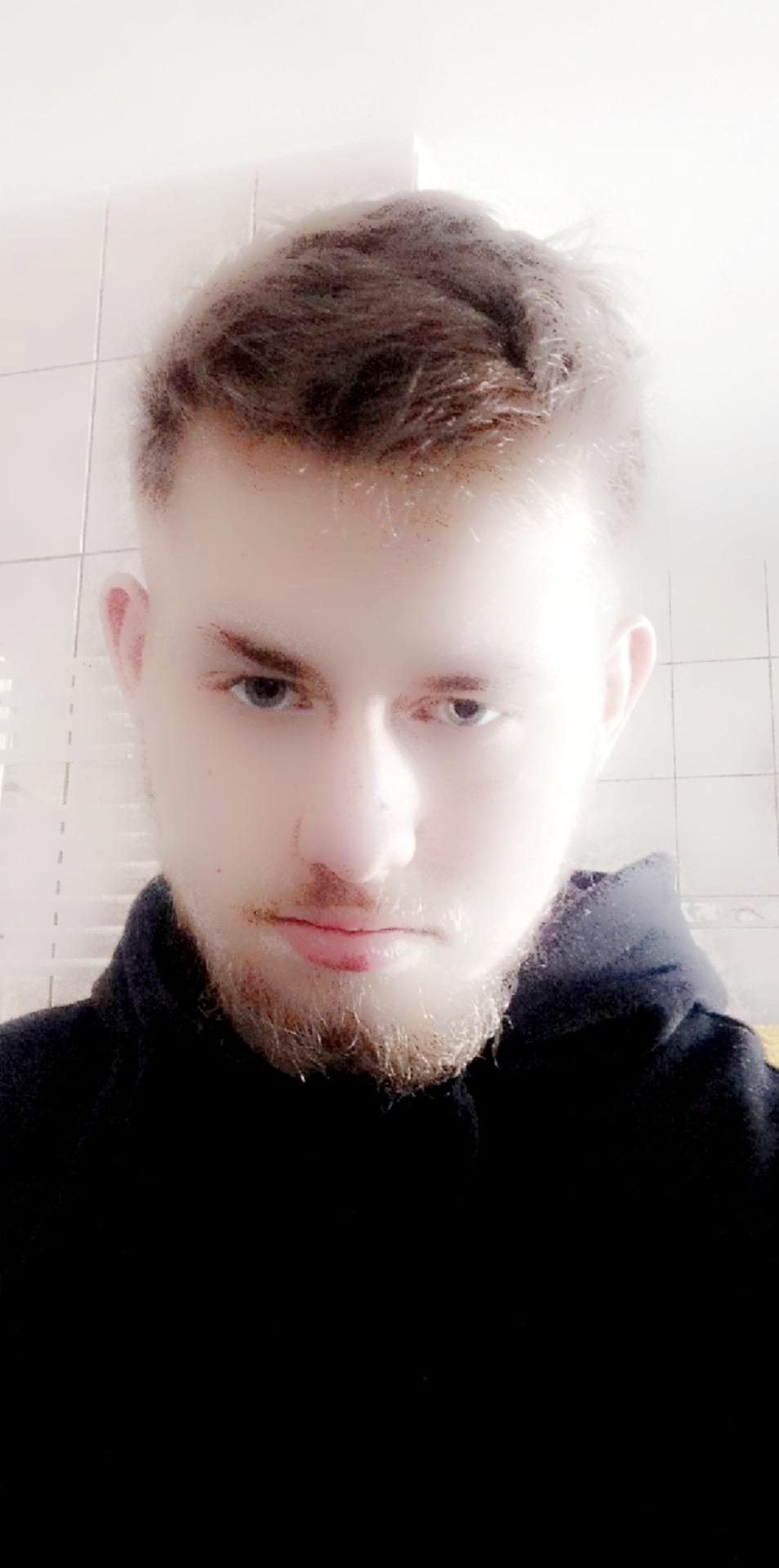 Portret młodego mężczyzny z lekkim zarostem, skierowany w dół, z intensywnym spojrzeniem na tle białych płytek. Ujęcie z góry, oświetlenie rozproszone.