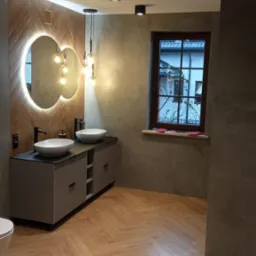 Elegancka łazienka po remoncie: drewnopodobna podłoga w jodełkę, podwójna umywalka nablatowa, okrągłe lustro z podświetleniem LED i nowoczesne oświetlenie sufitowe.